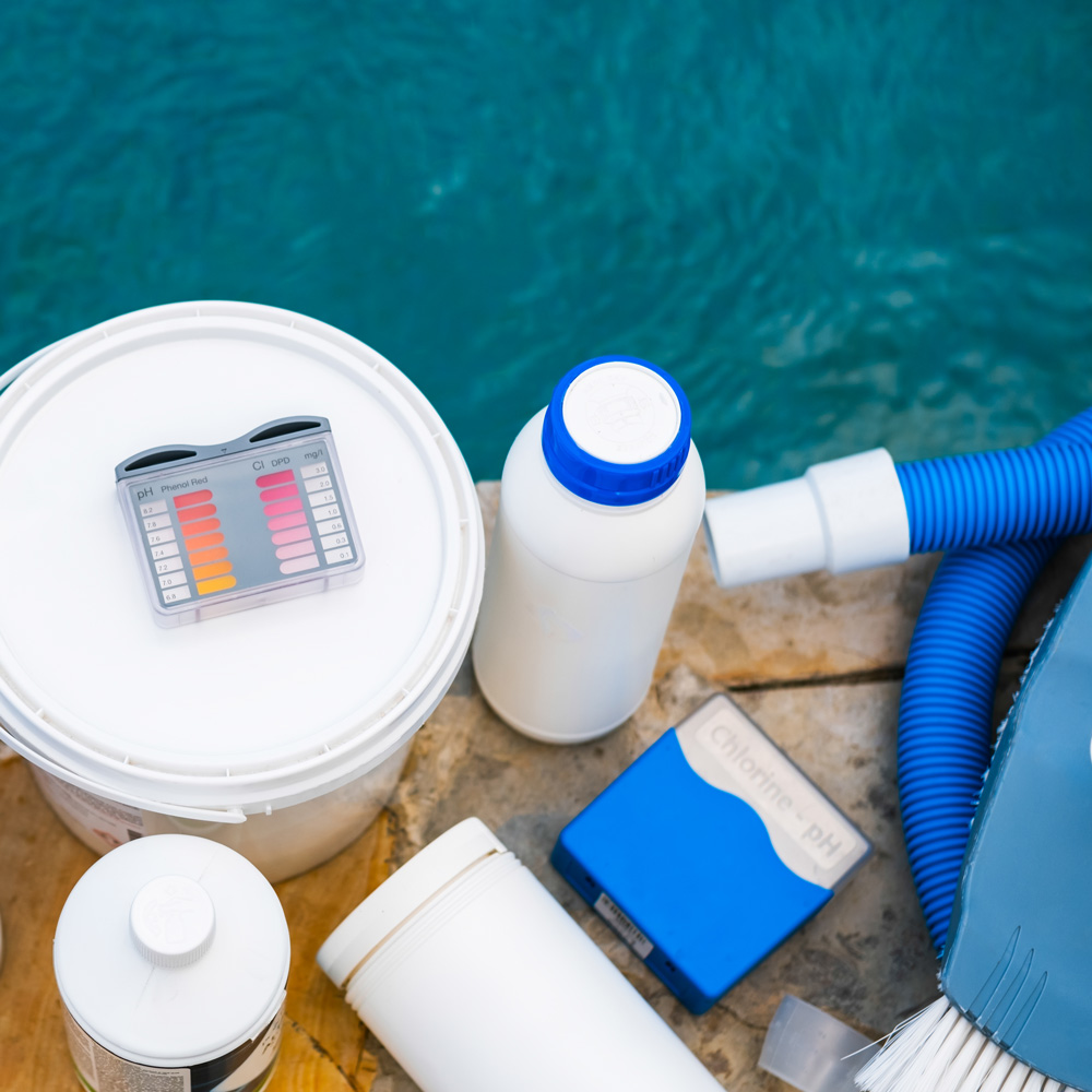 Pool-chemicals-and-equipment-by-the-side-of-pool Pool cleaning equipment, supplies, chemicals, and pH test kit sit on the patio by edge of pool